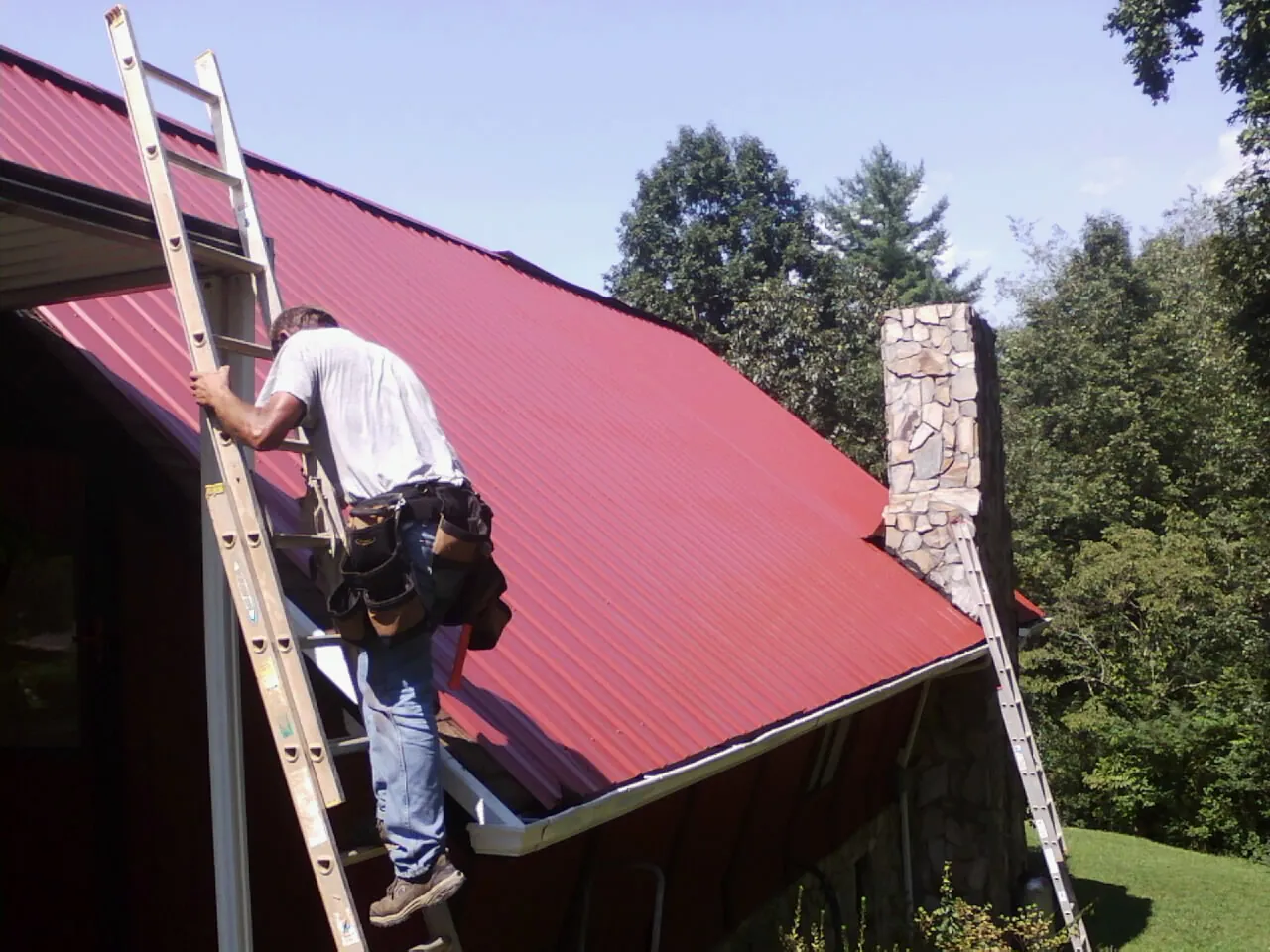 Roofer on a ladder beside a red metal roof and stone chimney — Soffit & Fascia Repair by Roofing Brothers in Glen Allen